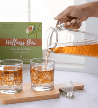 A hand pouring 100% all natural avocado leaf tea over ice, there are iced tea glasses and a wellness box filled herbal tea