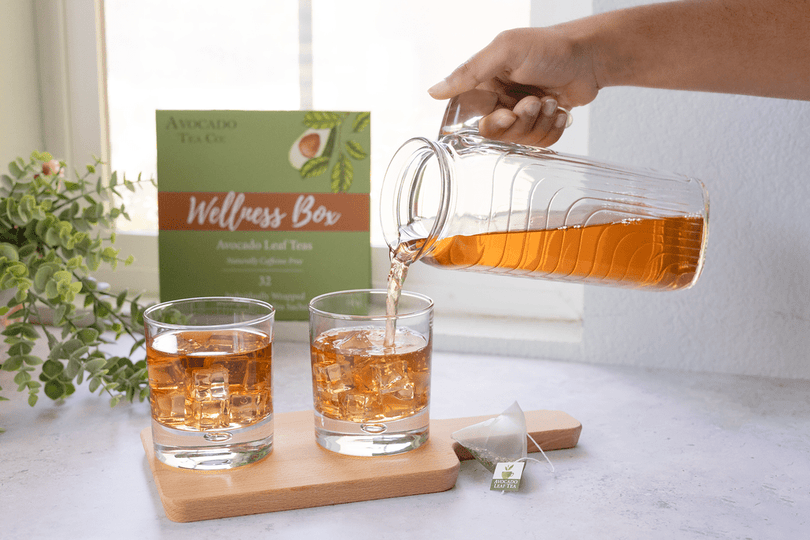 A hand pouring 100% all natural avocado leaf tea over ice, there are iced tea glasses and a wellness box filled herbal tea